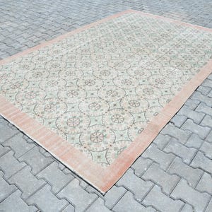 May include: A vintage rug with a cream background and a pink border. The rug has a repeating pattern of floral motifs in shades of green, pink, and white.