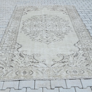 Può includere: Un tappeto rettangolare caratterizzato da una base color beige chiaro con un intricato motivo floreale e a medaglione in grigio scuro. Il design include un medaglione centrale circondato da motivi botanici decorati e un bordo dettagliato con elementi geometrici.