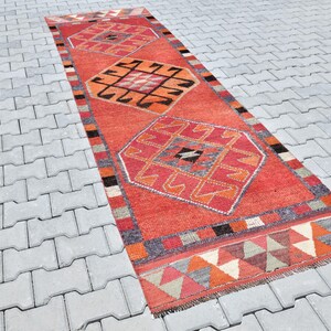 May include: A red, orange, and gray patterned rug with a geometric design. The rug is laid out on a gray concrete surface.