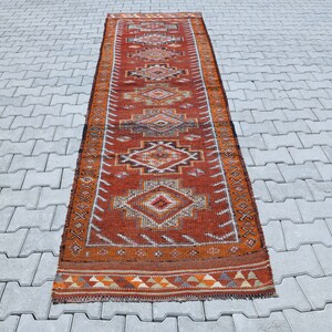 May include: A long, narrow, red and orange rug with a geometric pattern. The rug has a fringe on both ends.