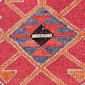 May include: Close-up of a red, orange, and purple patterned rug with a geometric design. The rug is made of woven fabric and has a textured surface.