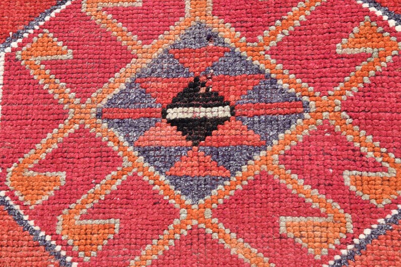 May include: Close-up of a red, orange, and purple patterned rug with a geometric design. The rug is made of woven fabric and has a textured surface.