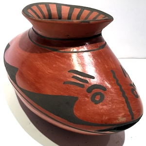 May include: A red clay pottery vase with black painted designs. The vase has a rounded body and a wide mouth. The design features a stylized fish with a black eye and a black line down the center.