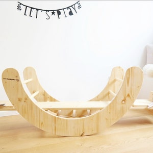 May include: A wooden rocking seesaw with a ladder on one side and a flat platform on the other. The seesaw is made of natural wood and has a rounded shape. The seesaw is in a room with white walls and a wooden floor.