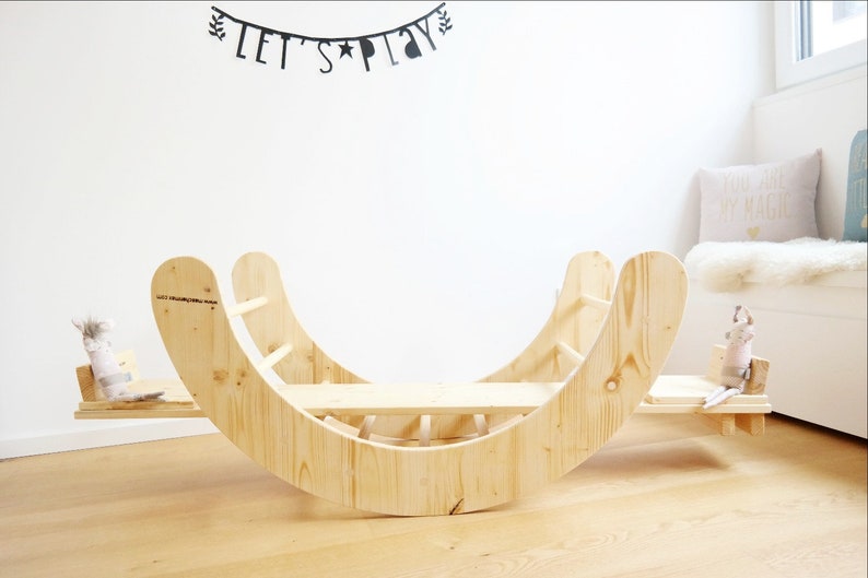 May include: A wooden rocking seesaw with a ladder on one side and a flat platform on the other. The seesaw is made of natural wood and has a rounded shape. The seesaw is in a room with white walls and a wooden floor.