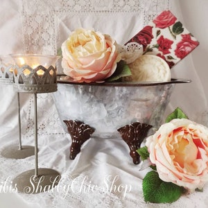 Könnte beinhalten: Eine weiße Metallwanne mit braunen Füßen, gefüllt mit einer weißen Seife, einem floral gemusterten Papier und rosa Rosen. Die Wanne steht auf einer weißen Spitzentischdecke mit zwei silbernen Kerzenständern.