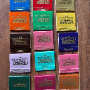 May include: A selection of incense matchboxes in various colours, each displaying "THE INCENSE MATCH" in gold. The boxes, in shades like blue, red, and green, are labelled with different scent names, such as 'sandalwood' and 'jasmine'.
