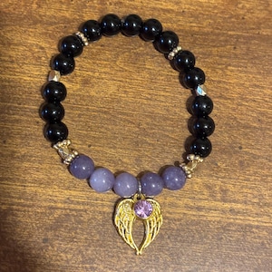May include: A beaded bracelet featuring smooth black spherical beads and a section of light purple beads. The bracelet includes gold-toned metal spacers and a gold-toned angel wing charm centered with a small, round, light purple gemstone.