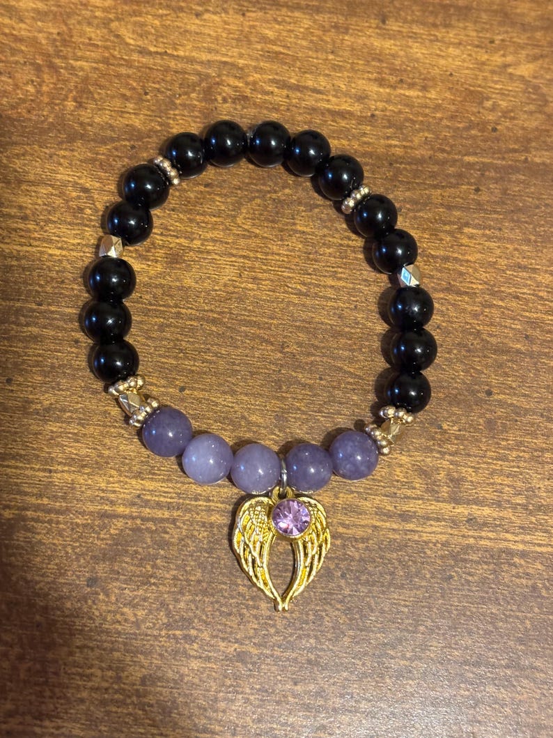 May include: A beaded bracelet featuring smooth black spherical beads and a section of light purple beads. The bracelet includes gold-toned metal spacers and a gold-toned angel wing charm centered with a small, round, light purple gemstone.