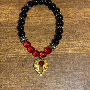 May include: A stretch bracelet featuring smooth black beads, four translucent red beads, and small metallic spacers. A gold-toned charm shaped like a pair of angel wings with a central red gemstone hangs from the bottom of the bracelet.