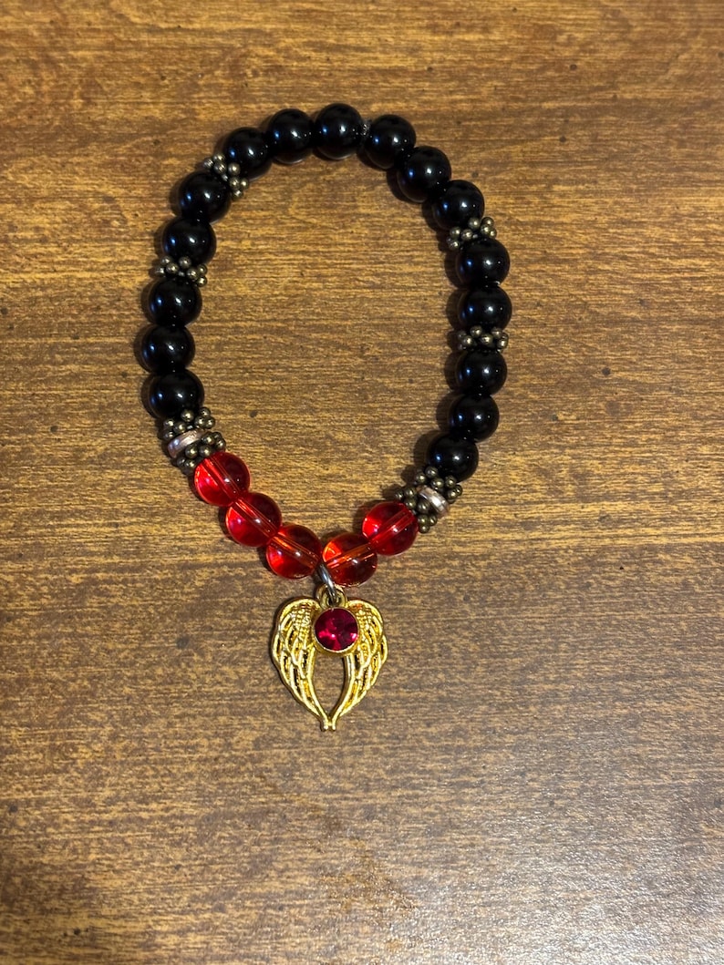 May include: A stretch bracelet featuring smooth black beads, four translucent red beads, and small metallic spacers. A gold-toned charm shaped like a pair of angel wings with a central red gemstone hangs from the bottom of the bracelet.