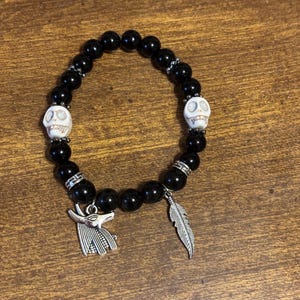 May include: Black beaded bracelet with two white skull charms, a silver ankh charm, and a silver feather charm.