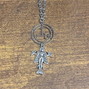May include: Silver-toned pendant necklace featuring a circular emblem with a cross-like design, connected to a detailed figure with wings and outstretched arms. The necklace hangs on a silver chain, set against a wooden background.