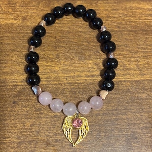 May include: A beaded stretch bracelet featuring glossy black spherical beads, five pale pink translucent beads, and small metallic heart-shaped spacers. A gold-toned angel wing charm with a central pink rhinestone hangs from the center.