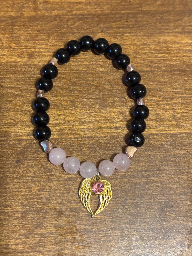 May include: A beaded stretch bracelet featuring glossy black spherical beads, five pale pink translucent beads, and small metallic heart-shaped spacers. A gold-toned angel wing charm with a central pink rhinestone hangs from the center.