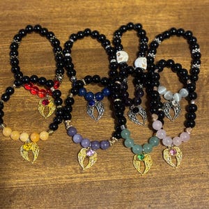 May include: A collection of beaded bracelets featuring black round beads, interspersed with colored gemstone beads and silver-toned angel wing charms. Each charm is accented with a small, round, colored rhinestone at the center.