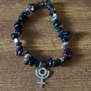 May include: A black, gray, and red gemstone bracelet with a silver charm featuring the astrological symbol for Uranus.