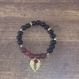 May include: A beaded bracelet featuring smooth black spherical beads and a section of deep red beads. The bracelet includes small gold-toned spacers and a gold-toned angel wing charm centered with a small, round, red gemstone accent.