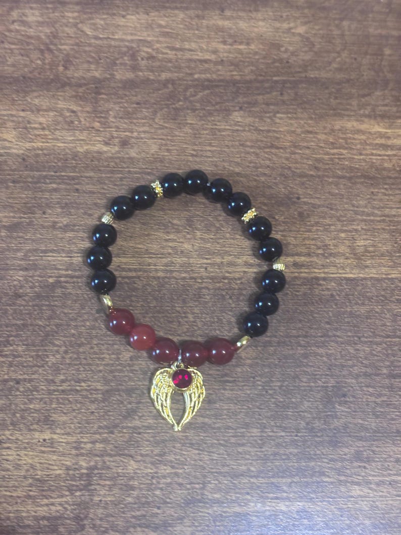 May include: A beaded bracelet featuring smooth black spherical beads and a section of deep red beads. The bracelet includes small gold-toned spacers and a gold-toned angel wing charm centered with a small, round, red gemstone accent.