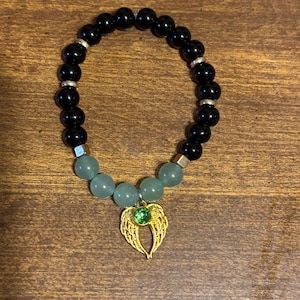 May include: A stretch bracelet featuring smooth black spherical beads, four pale green translucent beads, and small silver-toned spacers. A gold-toned metal charm shaped like angel wings with a central round green rhinestone is attached.