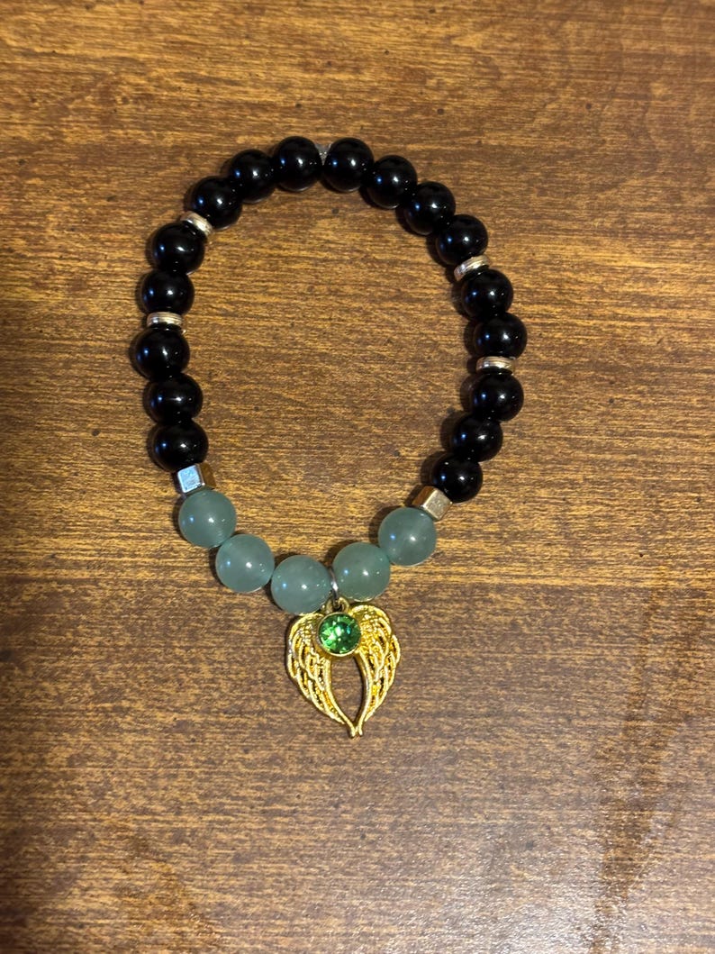May include: A stretch bracelet featuring smooth black spherical beads, four pale green translucent beads, and small silver-toned spacers. A gold-toned metal charm shaped like angel wings with a central round green rhinestone is attached.