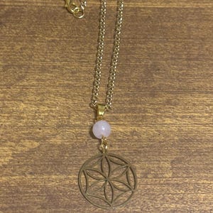 May include: Gold-colored necklace with a delicate chain. A light pink gemstone bead is suspended above a circular pendant with a floral design. The necklace is displayed on a wooden surface.