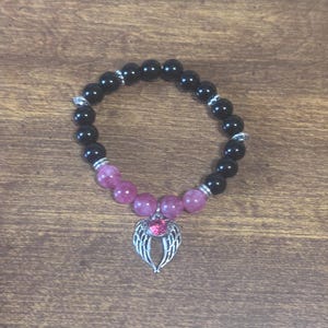 May include: A beaded bracelet featuring smooth black round beads and a section of translucent pink beads. The bracelet includes silver-toned metal spacers and a central silver-toned charm shaped like a pair of wings with a pink stone.