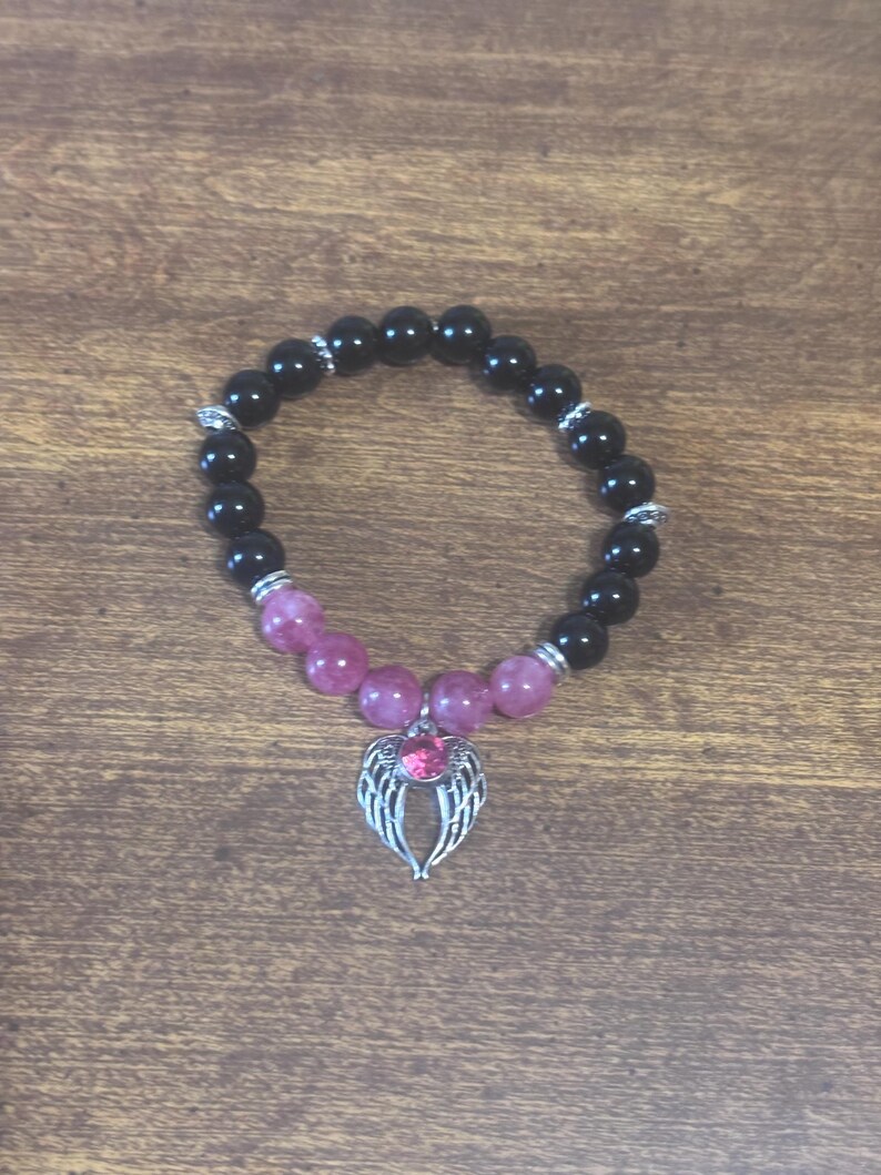 May include: A beaded bracelet featuring smooth black round beads and a section of translucent pink beads. The bracelet includes silver-toned metal spacers and a central silver-toned charm shaped like a pair of wings with a pink stone.