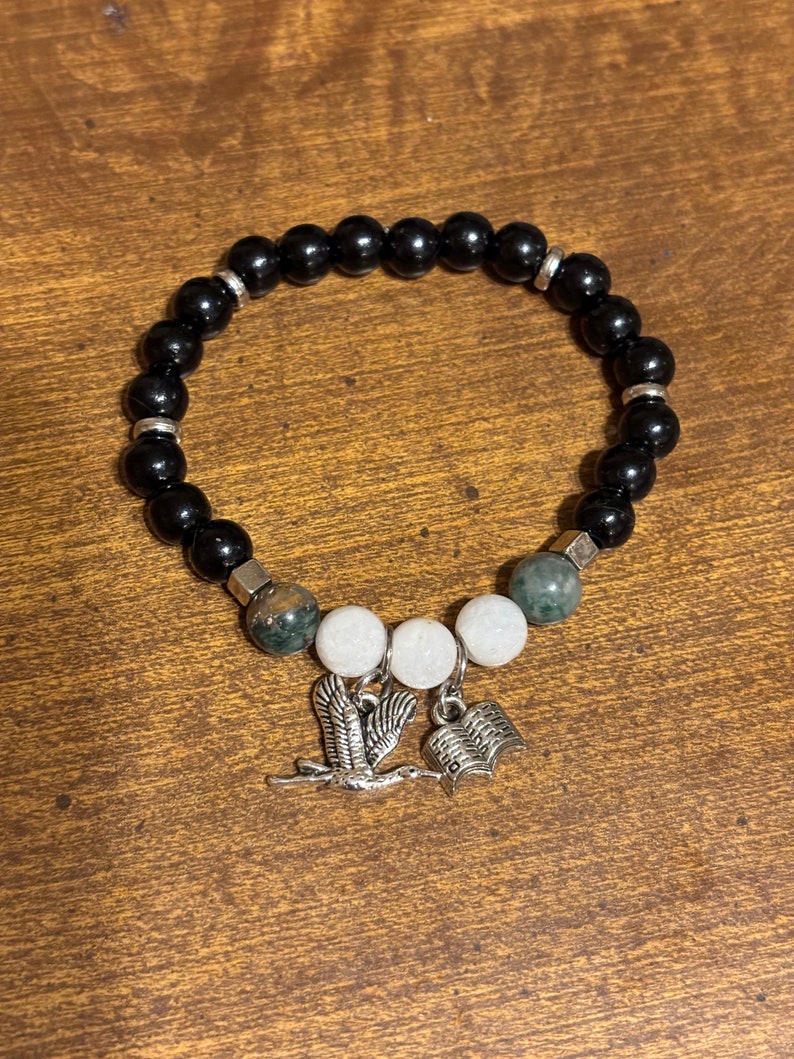 May include: Black beaded bracelet with green and white beads and silver charms of a bird and an open book.