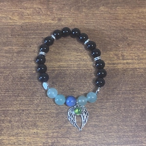 May include: A beaded bracelet featuring smooth black spherical beads, four pale green beads, and one central blue bead. The bracelet includes silver-toned spacers and a silver-toned angel wing charm with a small, round, bright green stone.