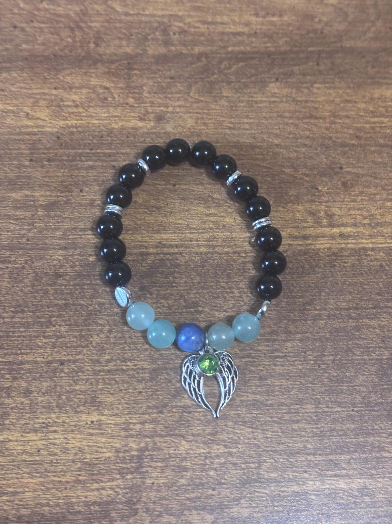 May include: A beaded bracelet featuring smooth black spherical beads, four pale green beads, and one central blue bead. The bracelet includes silver-toned spacers and a silver-toned angel wing charm with a small, round, bright green stone.