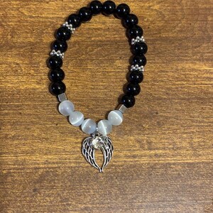 May include: A beaded bracelet featuring smooth black spherical beads, four translucent white cat's eye beads, and silver-toned spacers. A silver-toned metal charm shaped like angel wings with a central clear rhinestone hangs from the center.