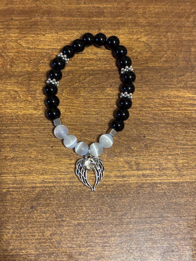 May include: A beaded bracelet featuring smooth black spherical beads, four translucent white cat's eye beads, and silver-toned spacers. A silver-toned metal charm shaped like angel wings with a central clear rhinestone hangs from the center.