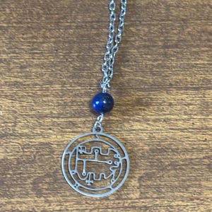 May include: A silver-colored pendant necklace with a circular charm and a blue bead. The charm has an intricate design within a circular frame. The necklace chain is made of small, linked pieces of metal.