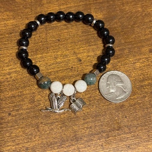 May include: Black beaded bracelet with silver accents and charms. The charms include a bird and an open book. The bracelet is shown next to a US quarter for size reference.