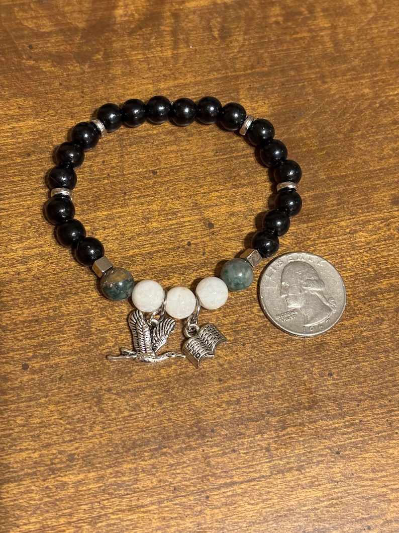 May include: Black beaded bracelet with silver accents and charms. The charms include a bird and an open book. The bracelet is shown next to a US quarter for size reference.