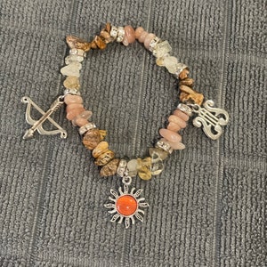 May include: A bracelet made with brown, pink, and white stones, with silver charms of a sun, an arrow and bow, and a treble clef.