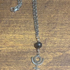 May include: Silver-toned necklace featuring a pendant with the astrological symbol for Pluto. The pendant hangs from a silver chain, with a dark gray bead accent. The necklace is displayed against a wooden background.