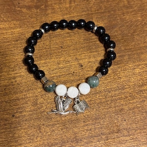 May include: Black beaded bracelet with white and green stone beads and silver charms of a bird and an open book.