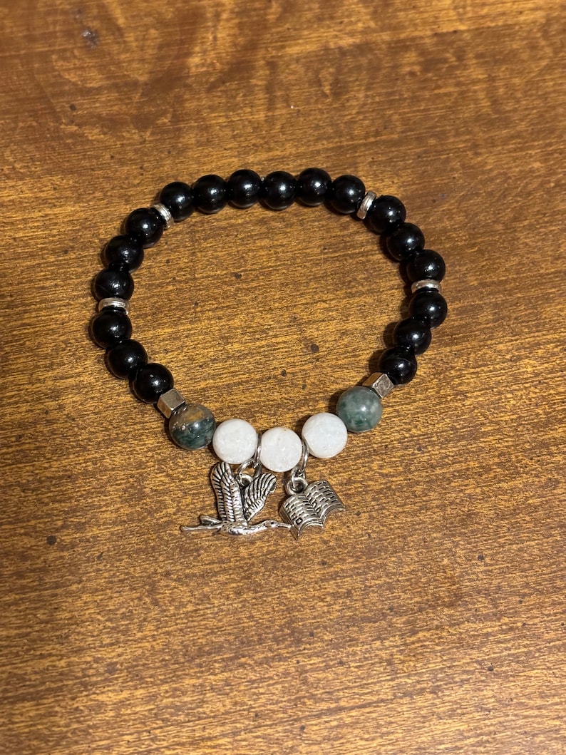 May include: Black beaded bracelet with white and green stone beads and silver charms of a bird and an open book.