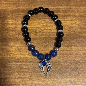 May include: A beaded bracelet featuring glossy black spherical beads, interspersed with silver-toned spacers and smaller blue beads. A silver-toned charm shaped like a pair of angel wings with a central blue circular stone hangs from the bracelet.