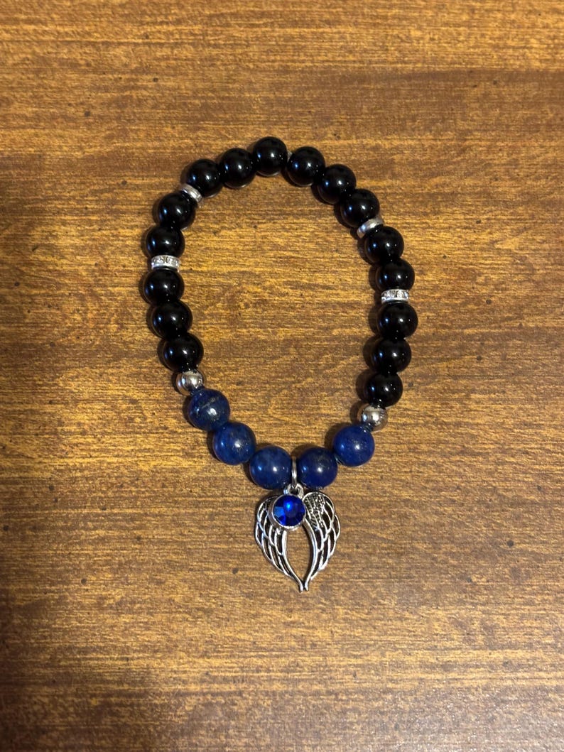 May include: A beaded bracelet featuring glossy black spherical beads, interspersed with silver-toned spacers and smaller blue beads. A silver-toned charm shaped like a pair of angel wings with a central blue circular stone hangs from the bracelet.