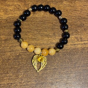 May include: A beaded bracelet featuring smooth black spherical beads and a section of translucent yellow-orange beads. The bracelet includes gold-toned metal spacers and a gold-colored charm shaped like angel wings with a central yellow gem.