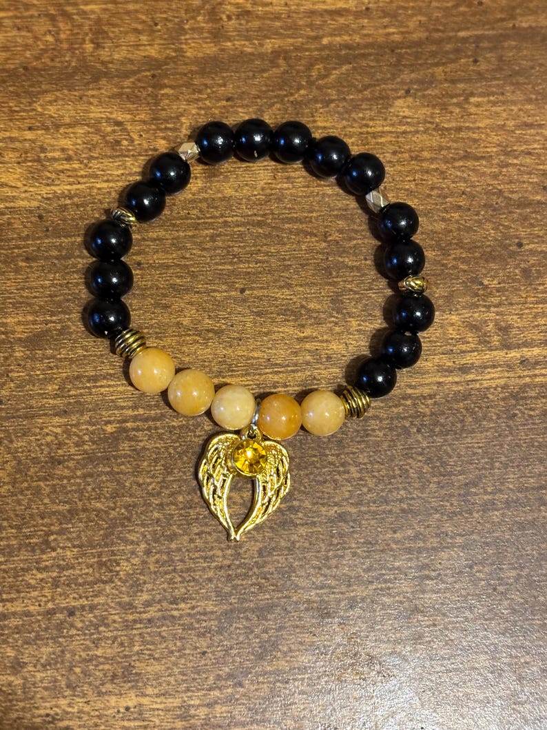 May include: A beaded bracelet featuring smooth black spherical beads and a section of translucent yellow-orange beads. The bracelet includes gold-toned metal spacers and a gold-colored charm shaped like angel wings with a central yellow gem.