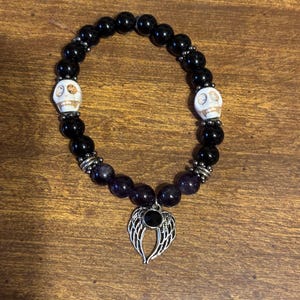 May include: A beaded stretch bracelet featuring smooth black round beads, two white skull-shaped beads with dark detailing, and silver-toned metal spacers. A silver-toned metal charm shaped like a pair of angel wings with a central black stone hangs from the bottom.