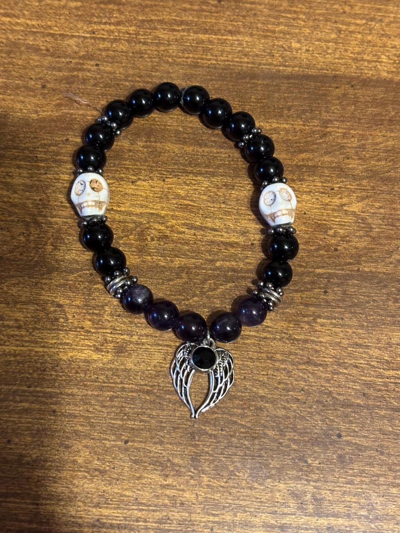 May include: A beaded stretch bracelet featuring smooth black round beads, two white skull-shaped beads with dark detailing, and silver-toned metal spacers. A silver-toned metal charm shaped like a pair of angel wings with a central black stone hangs from the bottom.