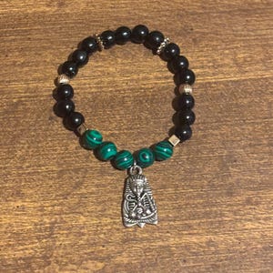 May include: Black beaded bracelet with malachite accents and a silver-tone pendant. The bracelet features a mix of black and green beads, with a detailed pendant of an Egyptian figure.