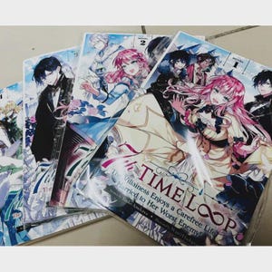 May include: A stack of manga novels with colorful illustrations. The covers feature anime-style characters with pink and blue hair. The title "TIME LOOP" is visible on one cover, along with the text "The Villainess Enjoys a Carefree Life Married to Her Worst Enemy!"