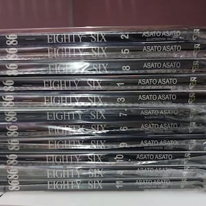 May include: A stack of 11 black and white manga volumes with the title "Eighty Six" and the name "Asato Asato" repeated on each volume. The volumes are numbered 1 through 11.
