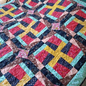 May include: A quilt with a geometric pattern in shades of red, yellow, blue, brown, and pink. The quilt is made of various fabrics with different patterns and textures.